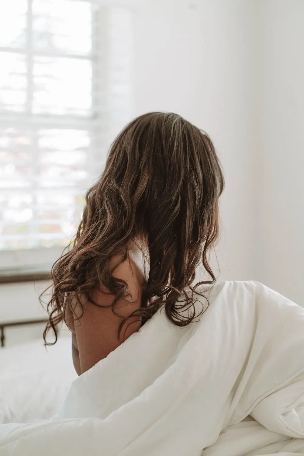 Une femme assise sur le bord du lit, de dos, le matin